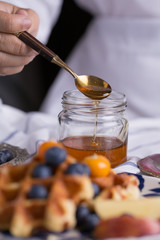Blueberry apple waffle with honey set on a dish with a chef's hand holding spoon for a jar of honey ready to serve.