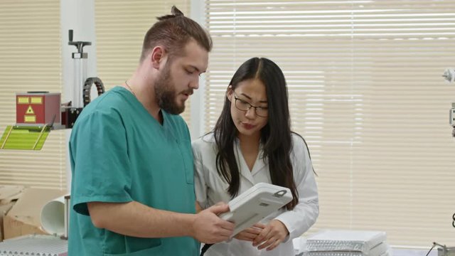 Engineer and young Asian woman in lab coat discussing process of making printed circuit board by machine