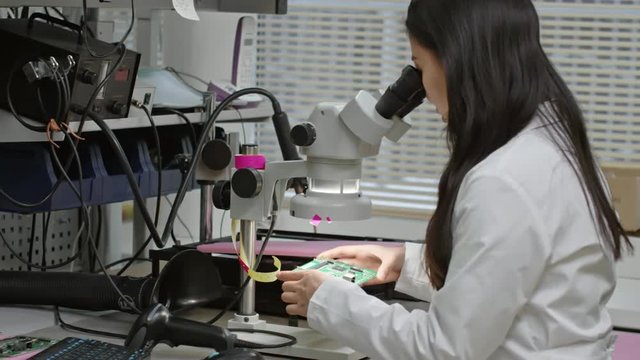 Female PCB inspector working in laboratory and checking printed circuit board under microscope
