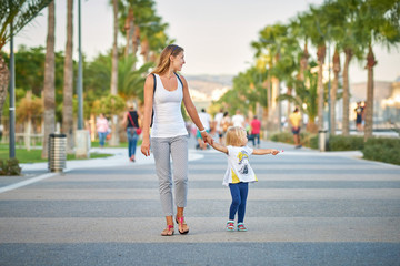 Young mother with her daughter walking at the promenade alley in the Molos Park of Limassol city, Cyprus - obrazy, fototapety, plakaty