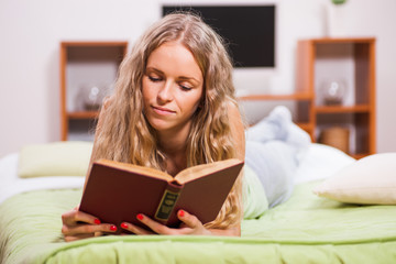 Young woman is lying in bed and reading book.