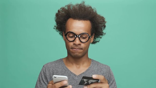 Focused young afro american man in eyeglasses using credit card and mobile phone for online shopping isolated on the green background