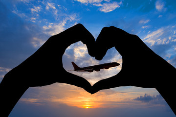 Silhouette of hands in the shape of a heart showing an airplane in flight, sky sunset background, air transport, travel and tourism