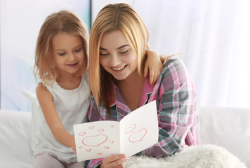 Beautiful young woman and her daughter sitting on bed with greeting card. Mother's day concept