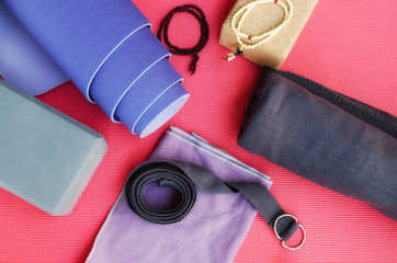 Yoga Accessories: rolled lilac exercise mat, cork block, grey strap, mala beads, towel and blanket on bright pink yoga mat background.