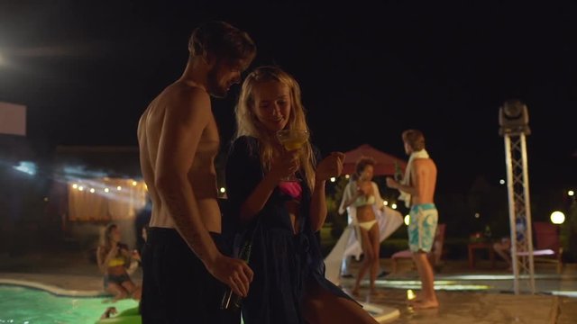 Topless Caucasian male with dark beard and beer dancing with blonde female with cocktail at night pool party flirting smiling. In slowmotion
