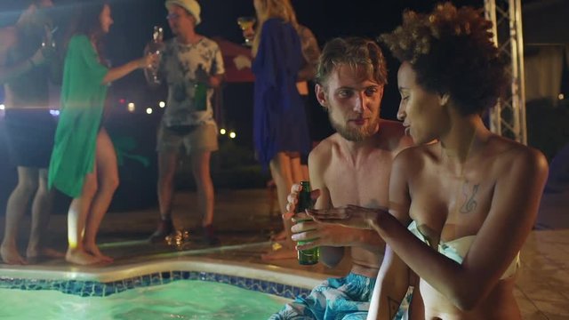 Caucasian topless male with light beard drinking beer and talking to African American female in white swimsuit laughing smiling in slowmotion at night pool party