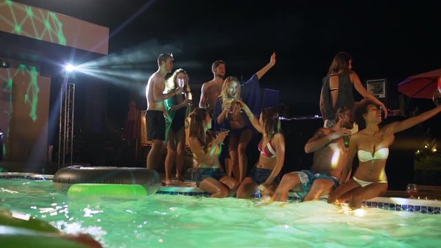 Group of beautiful males and females sitting standing at night pool party drinking cocktails at night pool party playing beachball smiling laughing talking in slowmotion