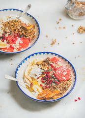 Healthy breakfast yogurt bowls with granola, grapefruit, banana, pomegranate, dried fruit, nuts, seeds over grey marble background, selective focus. Clean eating, vegetarian, vegan or dieting concept