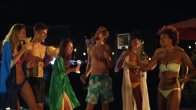 Group of cheerful men and women dancing in circle at night pool party with beers and cocktails in hands in slowmotion