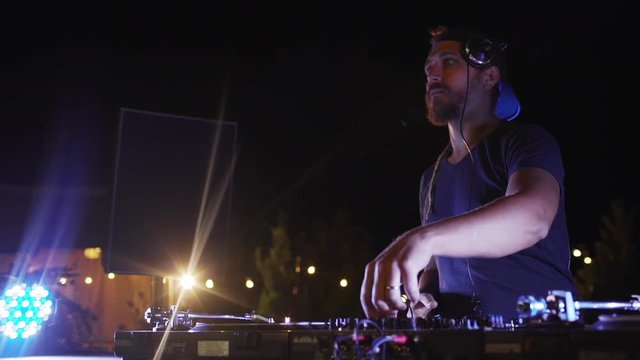 Bearded Caucasian DJ with cap and headphones playing music at outdoor pool party at night using mixer. In slowmotion