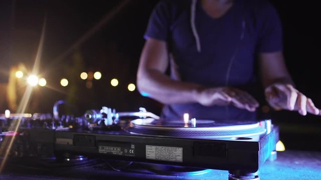 Caucasian DJ in blue t-shirt playing music at outdoor party at night pool party using mixer and vinyl disc. Close up in slowmotion