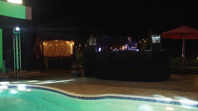 Caucasian bearded DJ in cap playing music at night pool party outdoors at night. In slowmotion