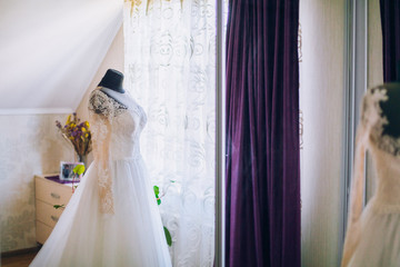 Beautiful blonde bride in robe posing near white wedding dress on mannequin