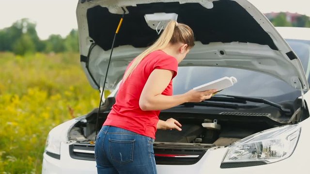Upset young woman looking at the car manual and trying to repair brakedown vehicle on the deserted road