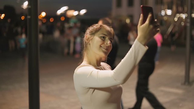 Portrait of elegant young woman making selfie on crowded street at night