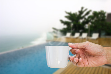 Hand holding hot coffee cup on blurred pool and beach background