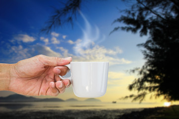 Hand holding hot coffee cup on blurred beach during sunrise background