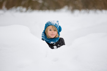 Fototapeta na wymiar Portrait of adorable little kid boy with long blond hair playing with snow outdoors. Child with blue scarf and hat walking and having fun on a windy winter day.