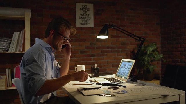 Young architect with light beard and blue shirt wearing glasses discussing on mobile phone project smiling making notes at table with lamp and laptop in slowmotion