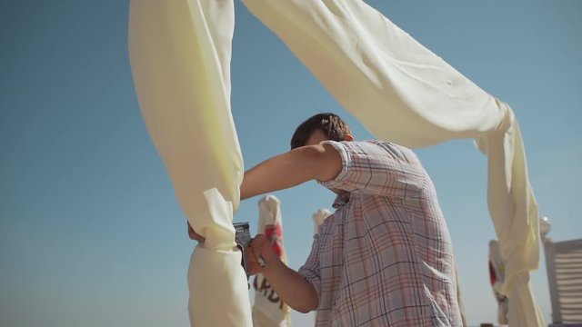 Caucasian decorators draping wedding arch near sea with staple gun in slowmotion