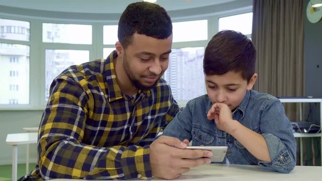 Little brunette boy flipping on father's smartphone. Attractive male kid using touchscreen of cellphone. Young african american father and his son watching images on the smartphone screen