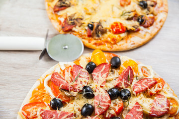 Pizza cutter together with two large pizzas with cheese olives and salami, mushrooms and tomatoes. On a wooden stand