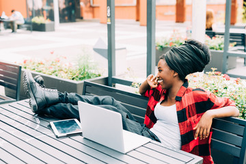 Young beautiful afro black woman sitting outdoor with legs on the table, relaxed, using computer and tablet, smiling - working, business, studying concept