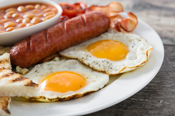 Traditional full English breakfast with fried eggs, sausages, beans, mushrooms, grilled tomatoes and bacon on wooden background

