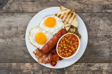 Traditional full English breakfast with fried eggs, sausages, beans, mushrooms, grilled tomatoes and bacon on wooden background
