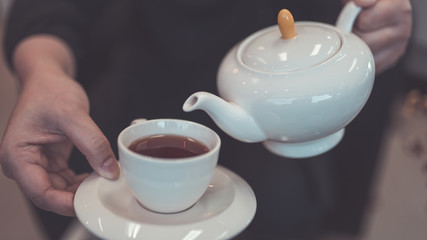 Hand holding a teapot pouring tea into a white cup on a wooden table. 