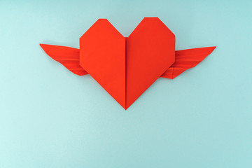 Red paper origami heart with wings on blue background .