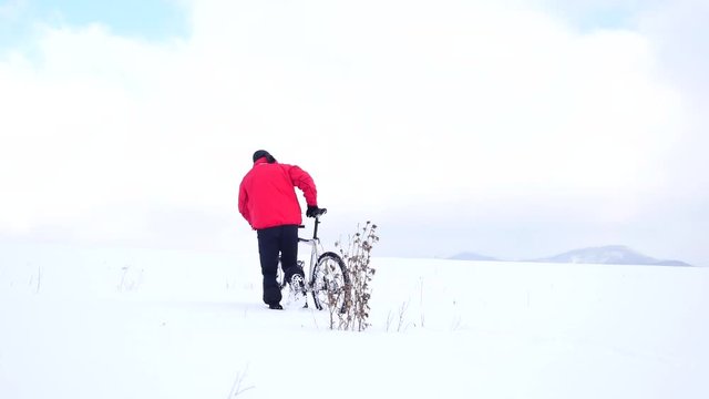 Man biker is pushing bike in deep snow at dry bush of thistle . Cloudy winter day with gentle wind and small snow flakes in the air. 