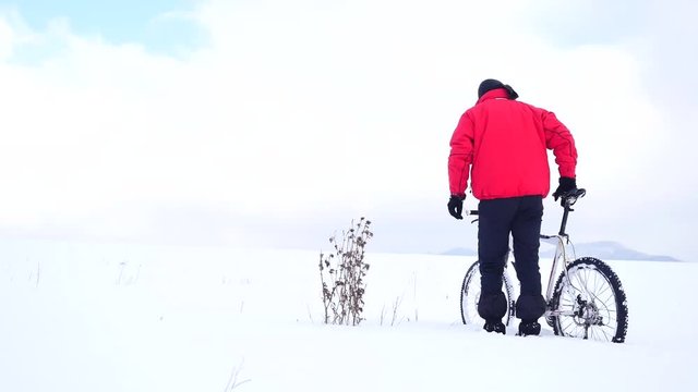 Man biker is pushing bike in deep snow at dry bush of thistle . Cloudy winter day with gentle wind and small snow flakes in the air. 
