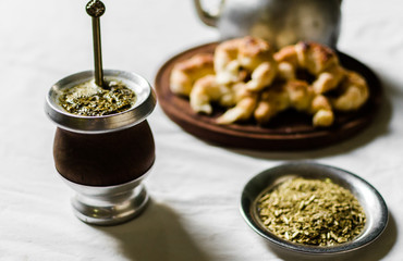 Close up of yerba mate tea in a calabash gourd, croissants on a wooden board, and kettle against white background. Selective focus