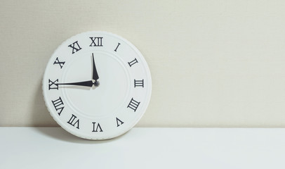 Closeup white clock for decorate show a quarter to twelve or 11:45 a.m. on white wood desk and cream wallpaper textured background with copy space
