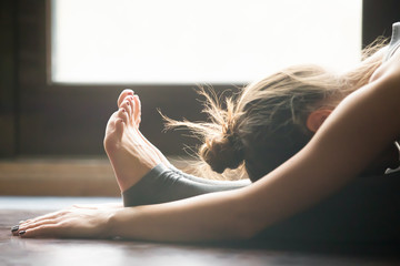 Young woman practicing yoga, sitting in Seated forward bend exercise, paschimottanasana pose, working out, wearing sportswear, grey pants, bra, indoor, home interior background, close up - obrazy, fototapety, plakaty