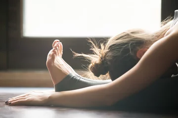Foto op Plexiglas Jonge vrouw die yoga beoefent, zittend in zittende voorwaartse buigoefening, paschimottanasana pose, trainen, sportkleding dragen, grijze broek, beha, binnen, interieur achtergrond, close-up © fizkes