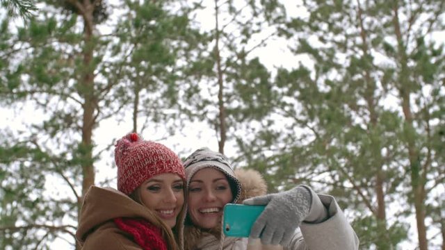 Tilt down of two joyous women photographing with smart phone in the forest at winter day