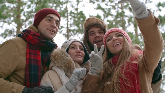 Group of young joyous men and women recording video with smart phone in winter forest. They smiling and telling something at camera