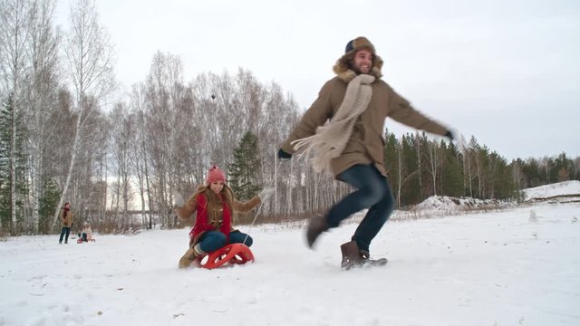 Man running on snow in park and pulling sleds with his laughing girlfriend