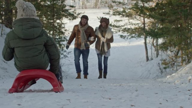 Slow motion of happy couple walking up snowy hill, and then running after sledding child 