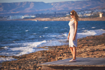Young woman with long hair standing at the edge of wooden pier and watching sunset at the rocky coast of Paphos, Cyprus