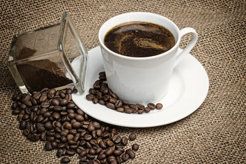 Higher angle view of the vintage photo of the white cup of coffee standing on the sackcloth. Around it are scattered coffee beans and ground coffee glass  
