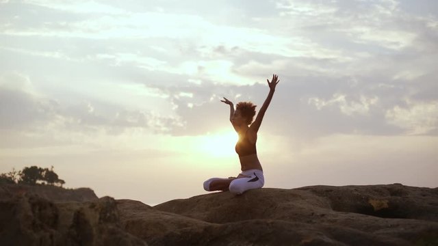 Young African American girl meditating in lotus posture on rocks and raising hands. Silhouette