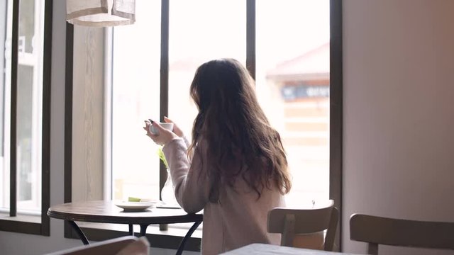 Back view of a young woman with long hair sitting and looking at the window while drinking coffee in cafe
