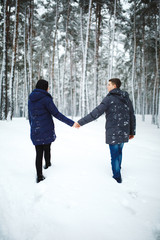 Fototapeta na wymiar Outdoor back view portrait of cheerful man and pretty girl enjoying the snowfall in winter forest. Young couple in love walking in wood. Christmas holidays