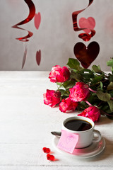 Closeup of cup of coffee with the bouquet of red roses of Valentine's day. Pink sticky note with the date February 14.