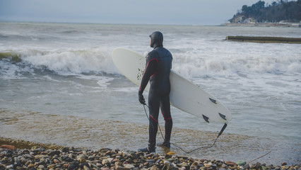 Зимний сёрфинг, сёрфер зимой в гидрокостюме идёт по пирсу в море с волнами
