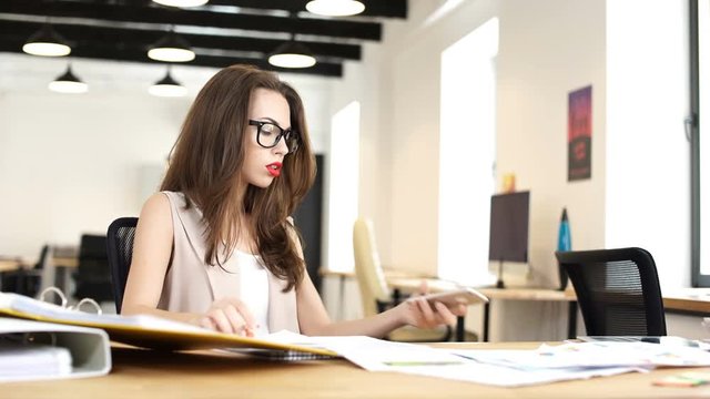 Young business woman making notes in documents and picking up mobile phone while sitting at her workplace